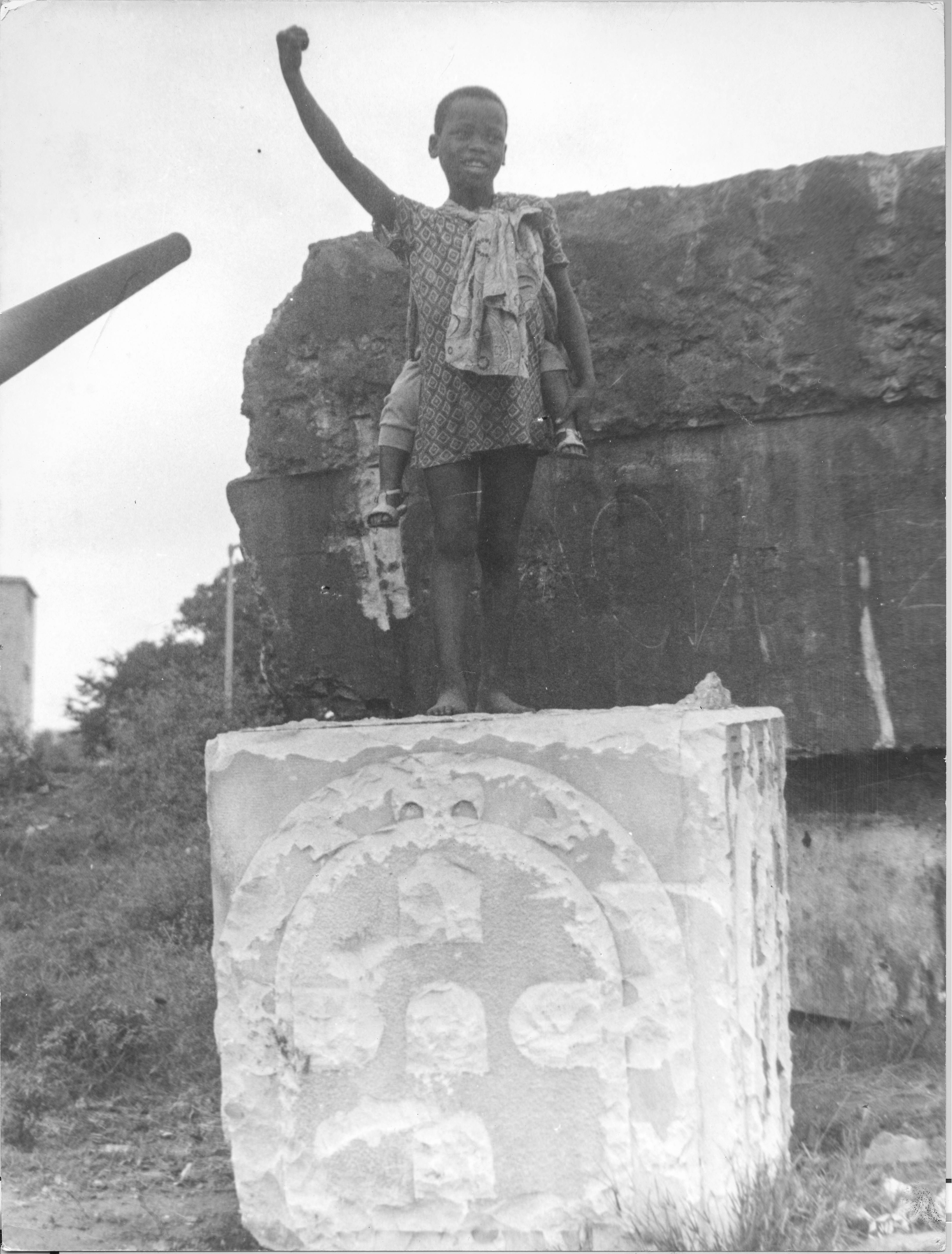Na imagem, uma criança com punho levantado, em cima de um monumento quebrado na ocasião da revolução com o Brasão de Portugal.